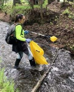 A woman wearing rubber boots, a gray backpack, work gloves, and a bright green shirt stands in a creek with a grabber and yellow plastic bag to pick up trash.